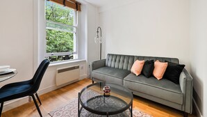 A modern living room with a grey sofa, black and pink cushions, a glass coffee table, a black chair, and a window with blinds. - Avari Apartments – Gray’s Collection (London)