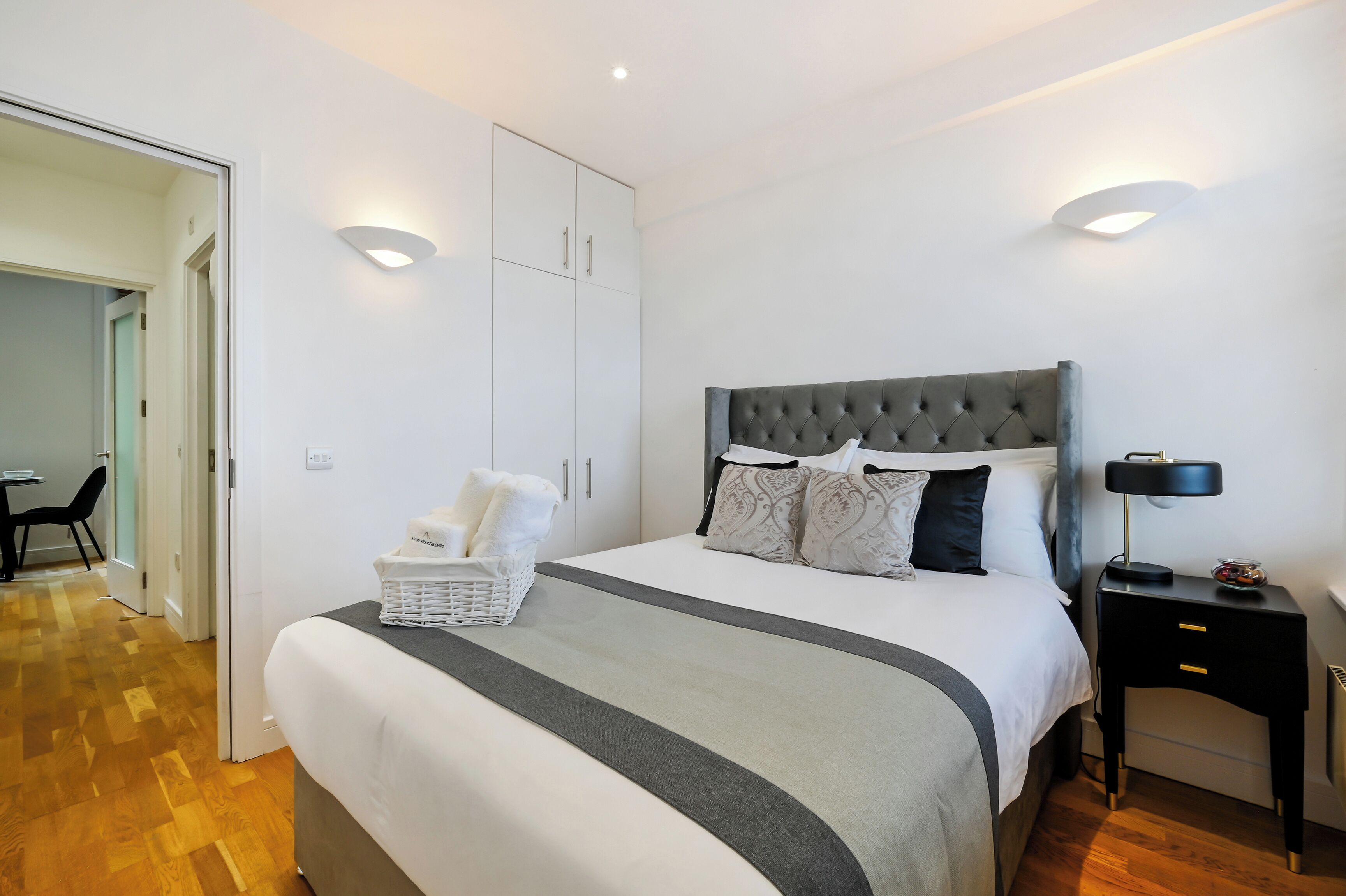 A neatly arranged bedroom with a large bed, a bedside table with a lamp, a wardrobe, and a chair in the background.