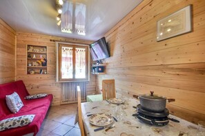 A cozy cabin interior with a red sofa, a dining table set with plates and glasses, a television mounted on the wall, and a window with curtains.