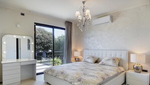 Ein Schlafzimmer mit einem großen Bett, einem Schminktisch mit Beleuchtung, einer Kristallleuchtre und einem Balkon mit Blick auf Bäume.