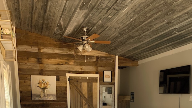 A bedroom with a bed, a ceiling fan, a wooden wall, a painting, a mirror, a TV, and a small table. - Charming luxury studio cottages with hints of rustic flare to help you RECHARGE (Woodville)