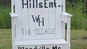A white wooden chair with text indicating 'Woodland HillsEnt., WH THE VILLAGE, Woodville, Ms'. - Charming luxury studio cottages with hints of rustic flare to help you RECHARGE (Woodville)