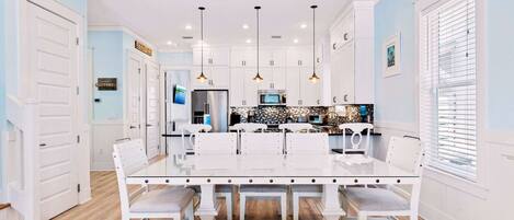 A modern kitchen with white cabinetry, a central island, pendant lighting, and a dining area with a white table and chairs.
