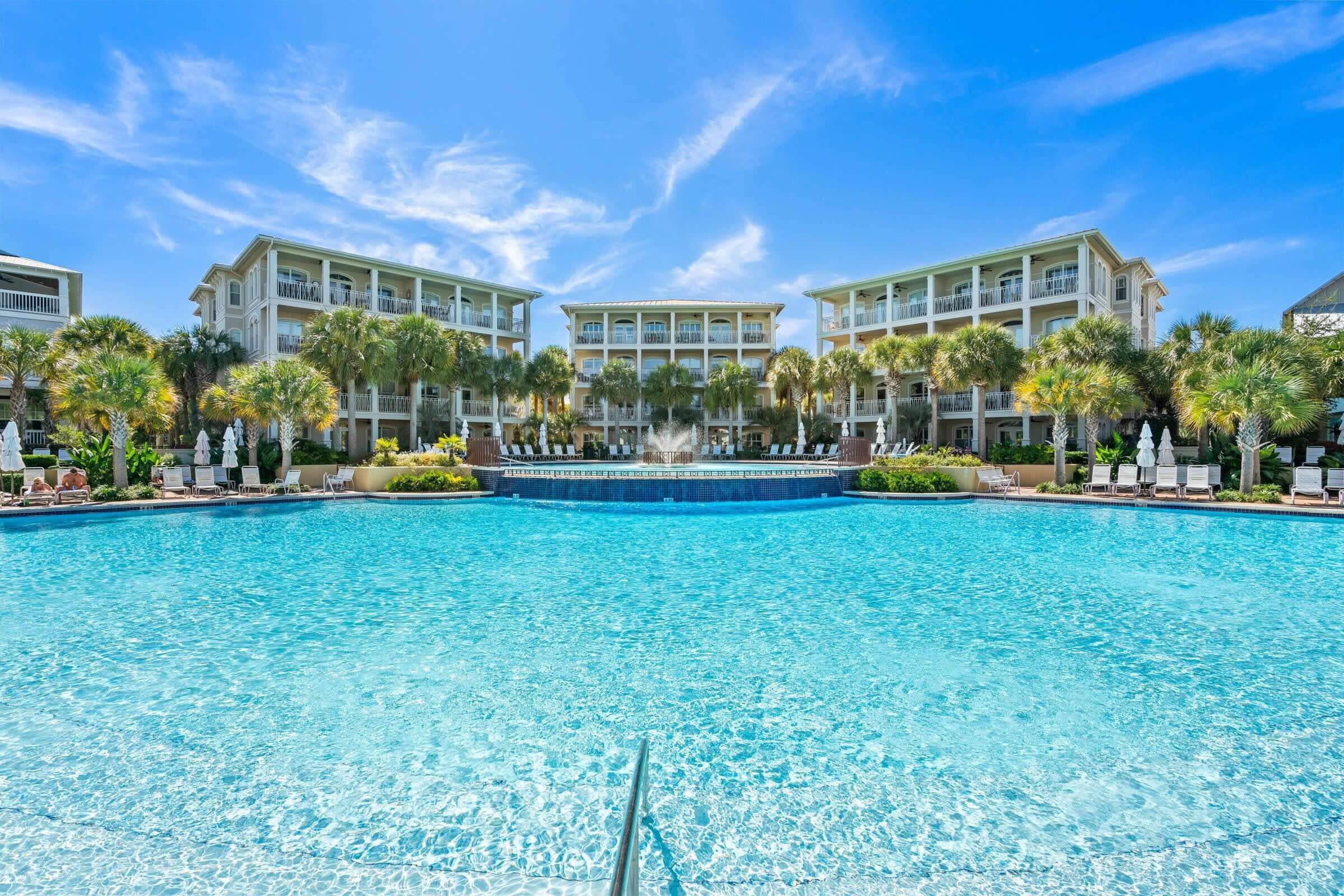 A large outdoor swimming pool with a fountain, surrounded by palm trees and lounge chairs, with a multi-story building in the background.