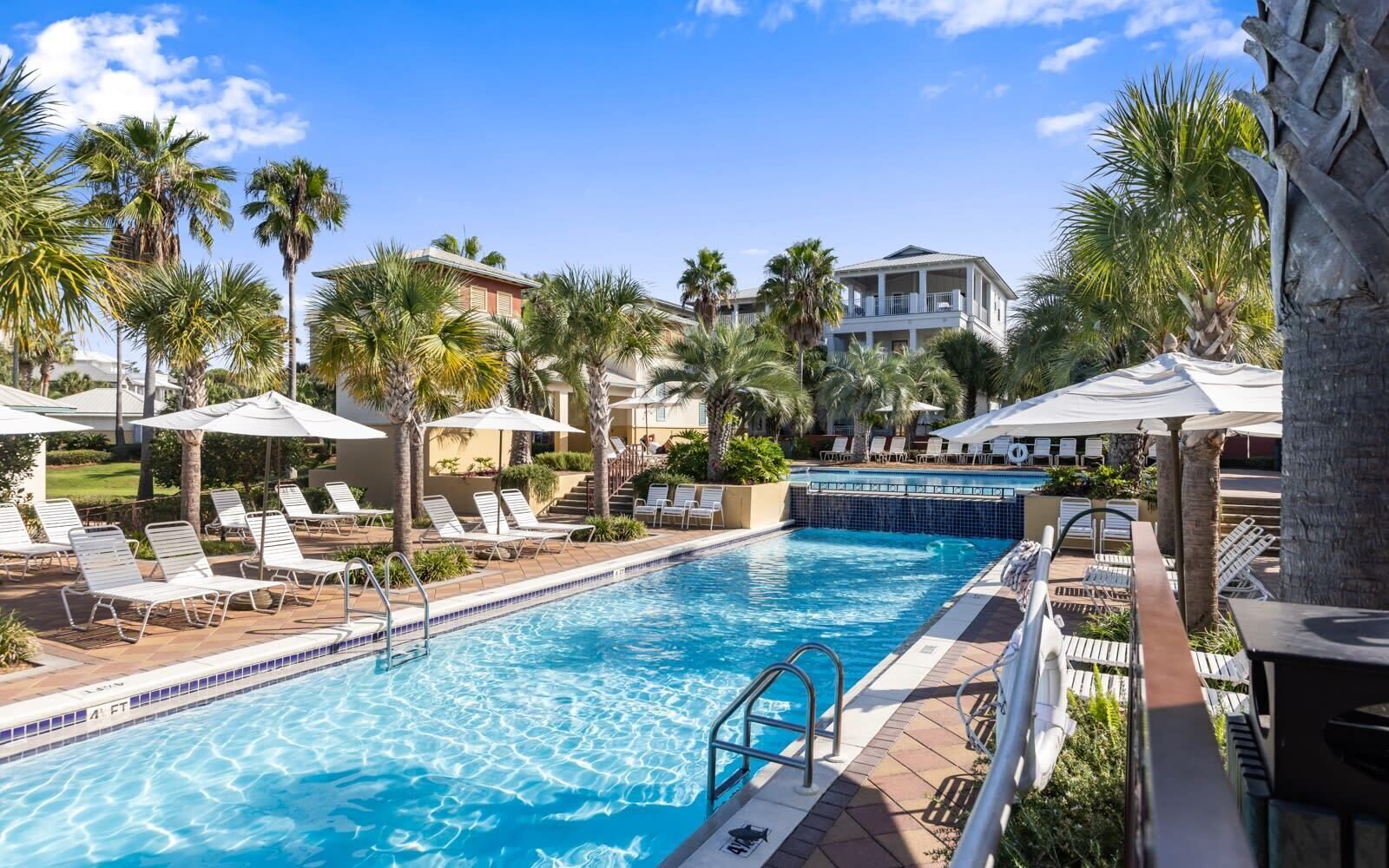 A swimming pool with lounge chairs and umbrellas, surrounded by palm trees and a building with a balcony.