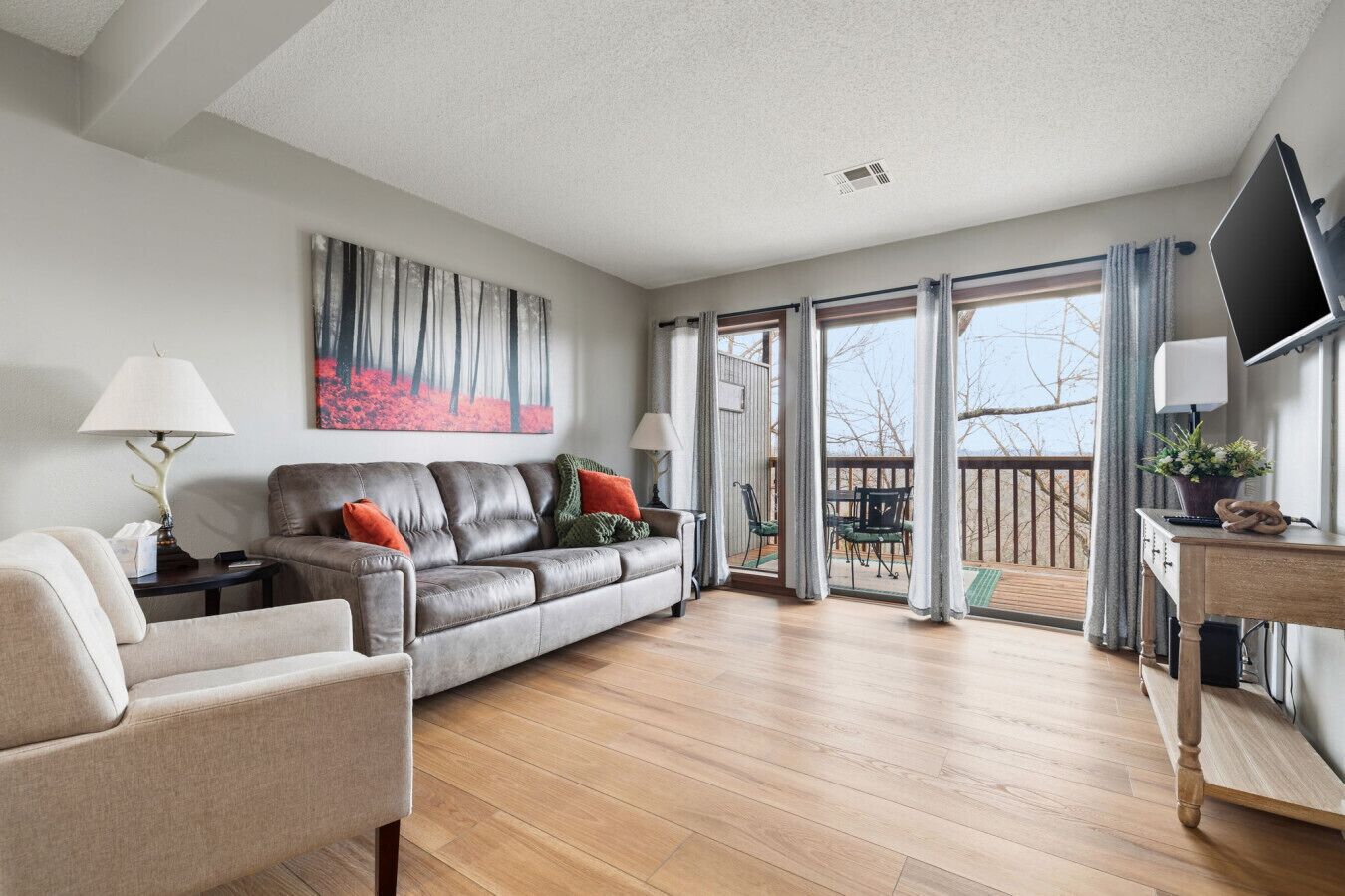Ein großzügiger Wohnraum mit grauem Sofa, beigem Sessel und einem Holztisch. Ein großes Gemälde ziert die Wand, und ein Fernseher ist an der Wand befestigt.