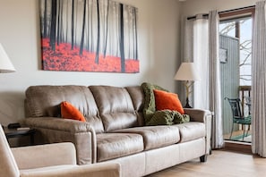 A living room with a leather sofa, two pillows, a painting, a lamp, and a view through a glass door.