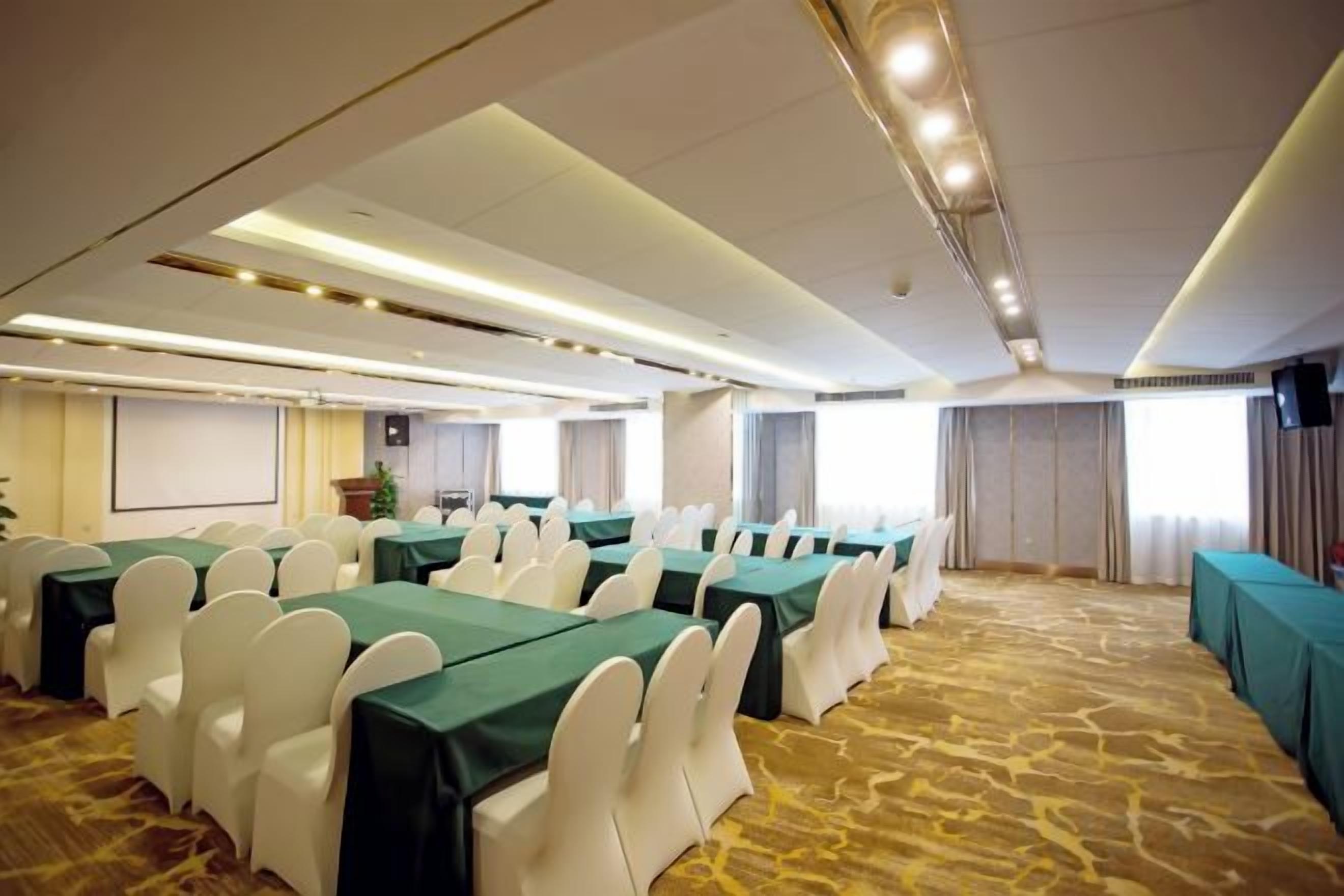 A conference room with rows of tables and chairs, a projector screen, and a carpeted floor.