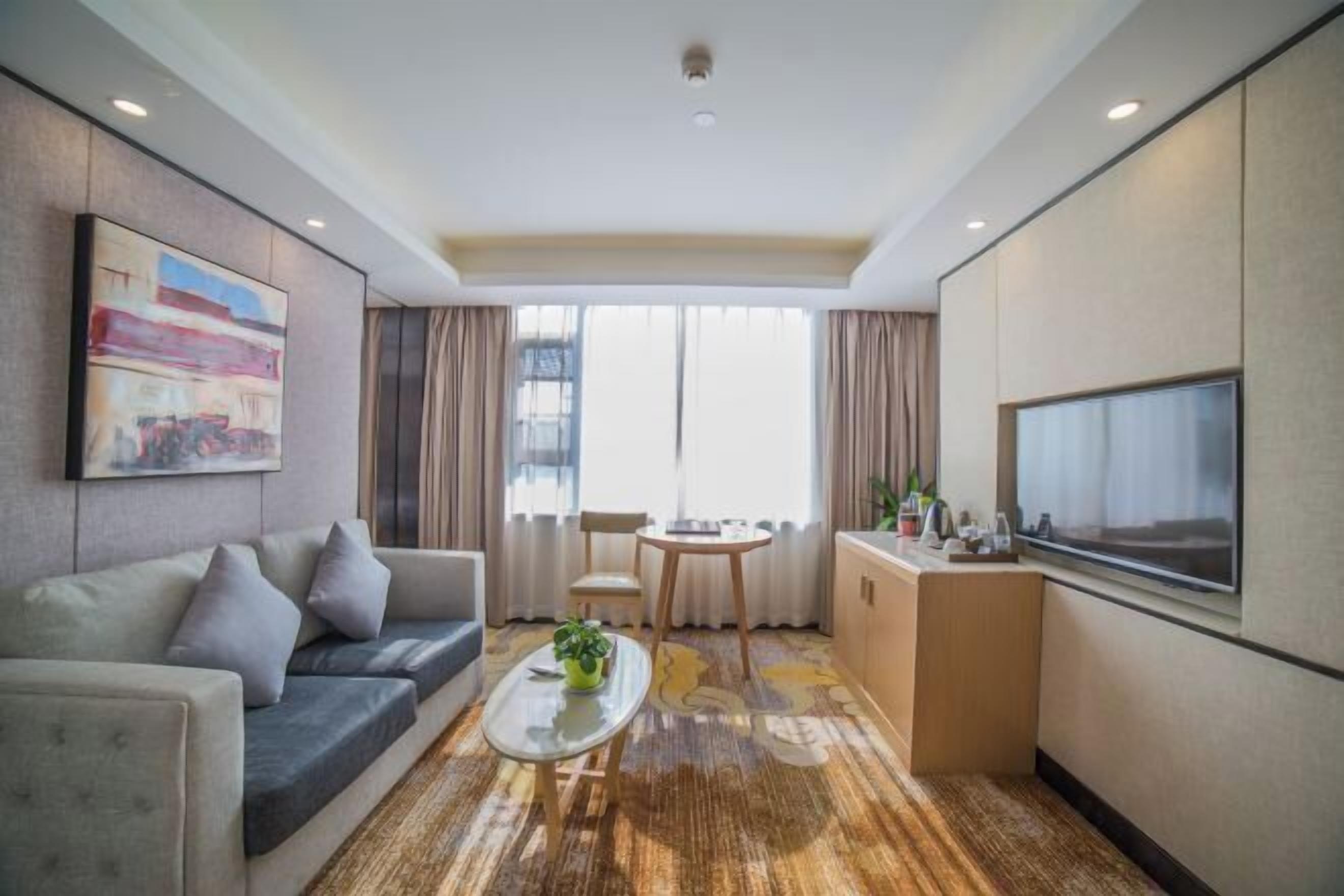 A hotel room with a sofa, a small table with a plant, a TV, a kitchenette, and a large window with curtains.