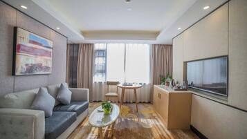 A hotel room with a sofa, a small table with a plant, a TV, a kitchenette, and a large window with curtains.