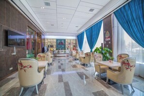 Een hotel lobby met een centraal zitgedeelte, een wandtelevisie en een groot raam met blauwe gordijnen.