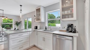 A modern kitchen with white cabinetry, a dishwasher, a coffee maker, and a large window offering a view of greenery.