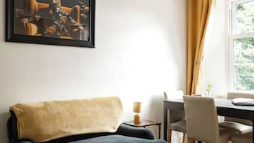 A living/dining room with a dark sofa, a dining table with chairs, a framed artwork on the wall, and a hanging light fixture.