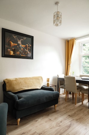 Una sala moderna con un sofá oscuro, una mesa de comedor con sillas, un cuadro de ciervos y una ventana con cortinas.
