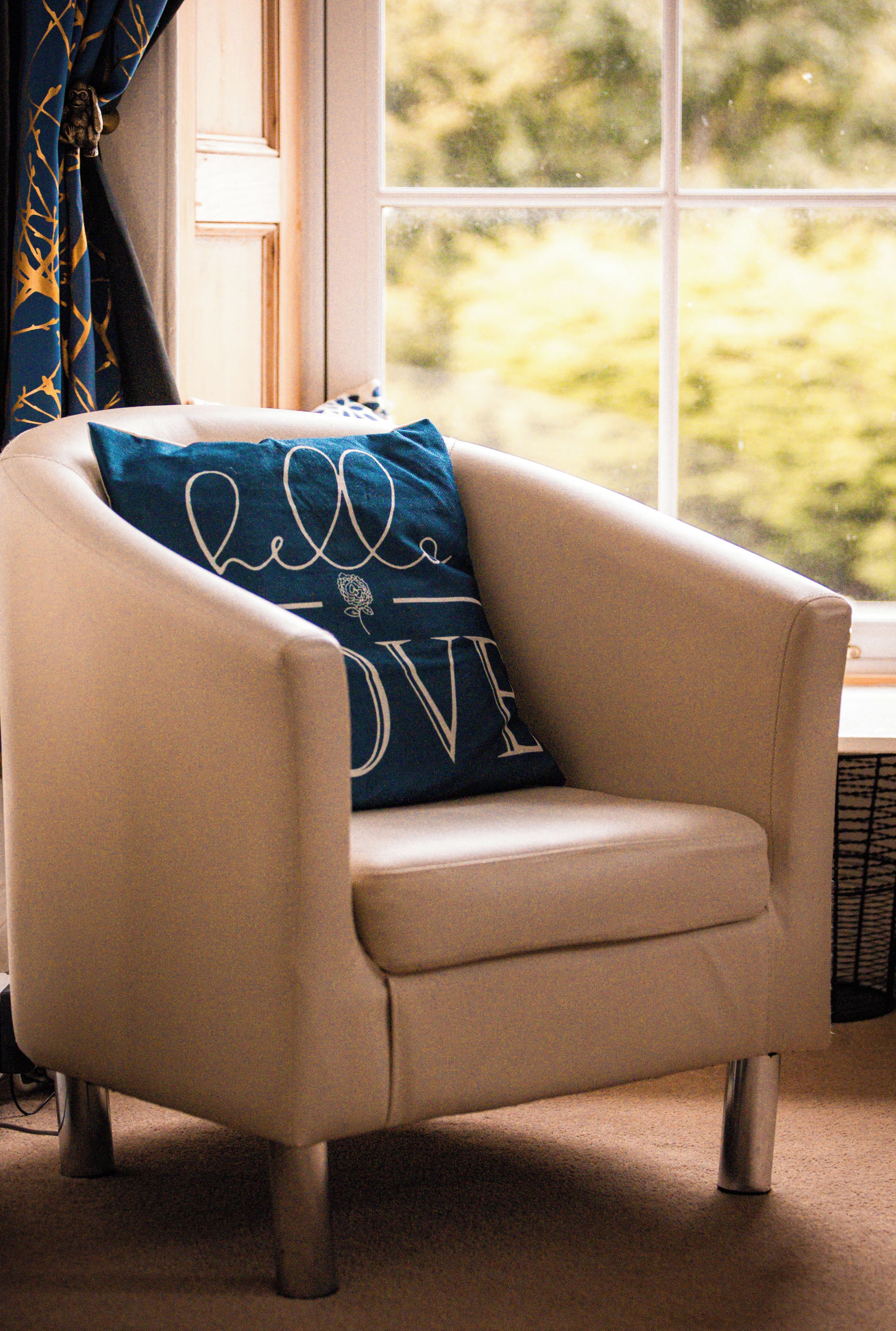 A beige armchair with a cushion that reads "hello LOVE" in white cursive letters, positioned in a room with a large window and a view of greenery outside.
