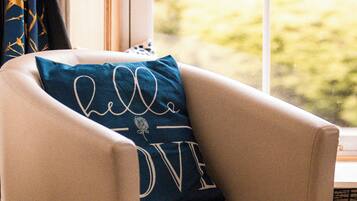 A beige armchair with a cushion that reads "hello LOVE" in white cursive letters, positioned in a room with a large window and a view of greenery outside.