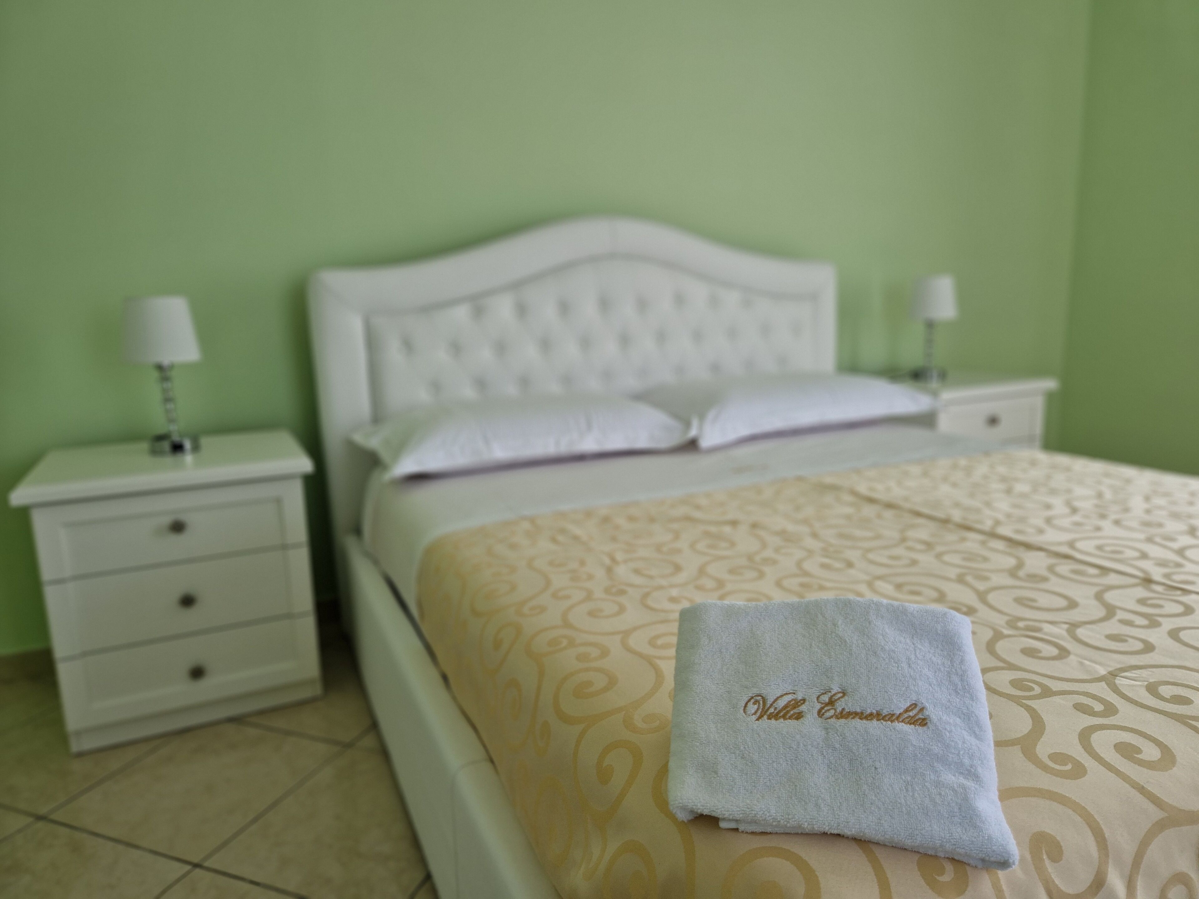A neatly made bed with a patterned bedspread and a towel with text, a white nightstand with a lamp, and a green wall.