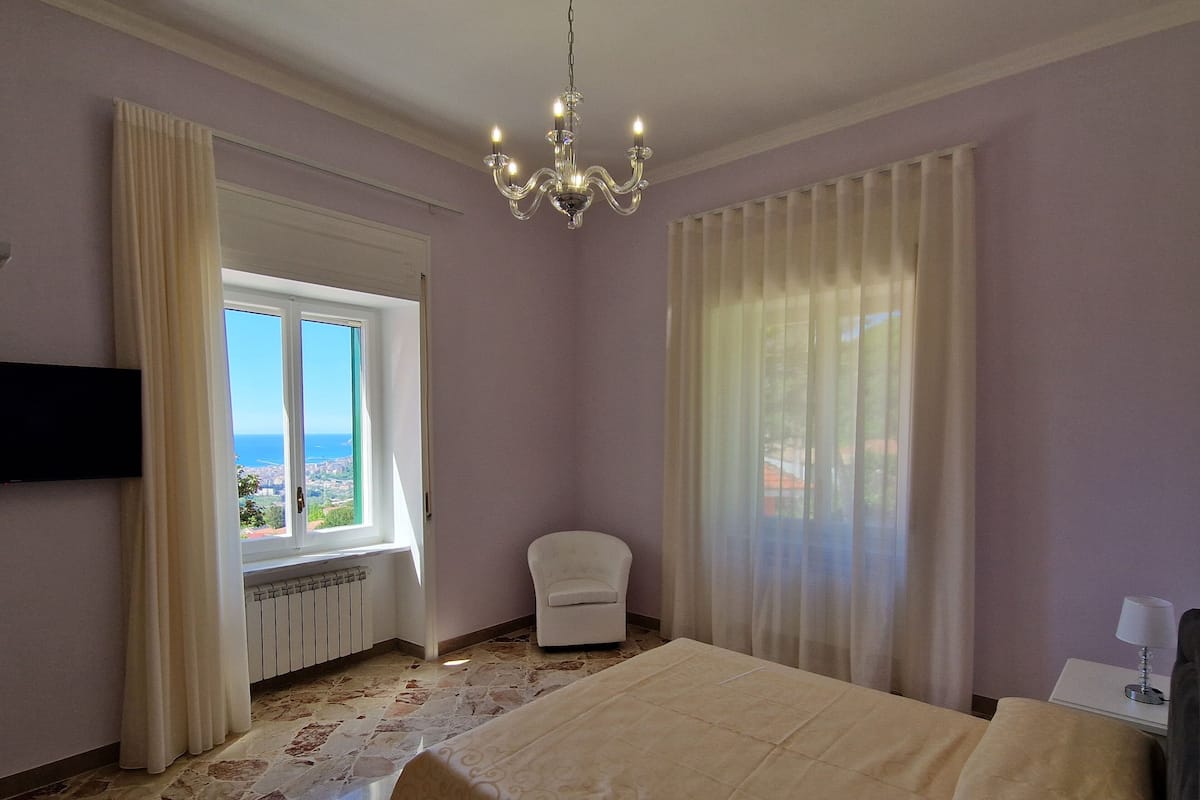 Ein Schlafzimmer mit Bett, Sessel, Fernseher, einem Fenster mit Aussicht und einer Kristallleuchter.