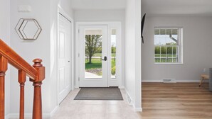 Een hal met een houten trap, een dubbele deur naar buiten en een raam met uitzicht op groen.
