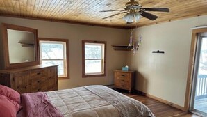 A bedroom with a wooden ceiling, a bed with a pink and white bedspread, a dresser, and two windows.
