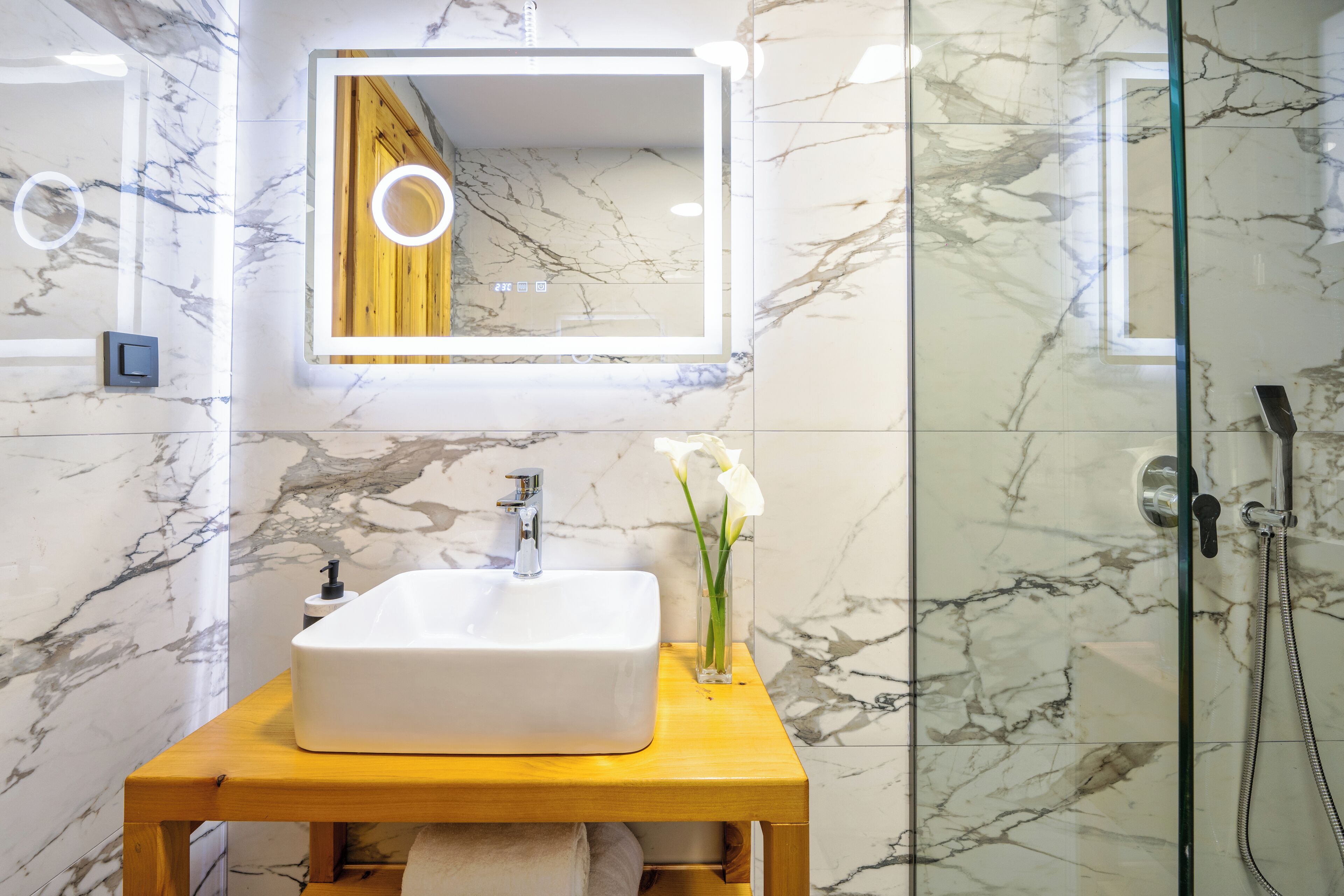 Un baño con pared de mármol, tocador de madera, lavamanos blanco, espejo con luz y cabina de ducha de vidrio.