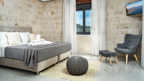 Un dormitorio con pared de piedra, cama con cabecera gris, sillón gris y otomana redonda.