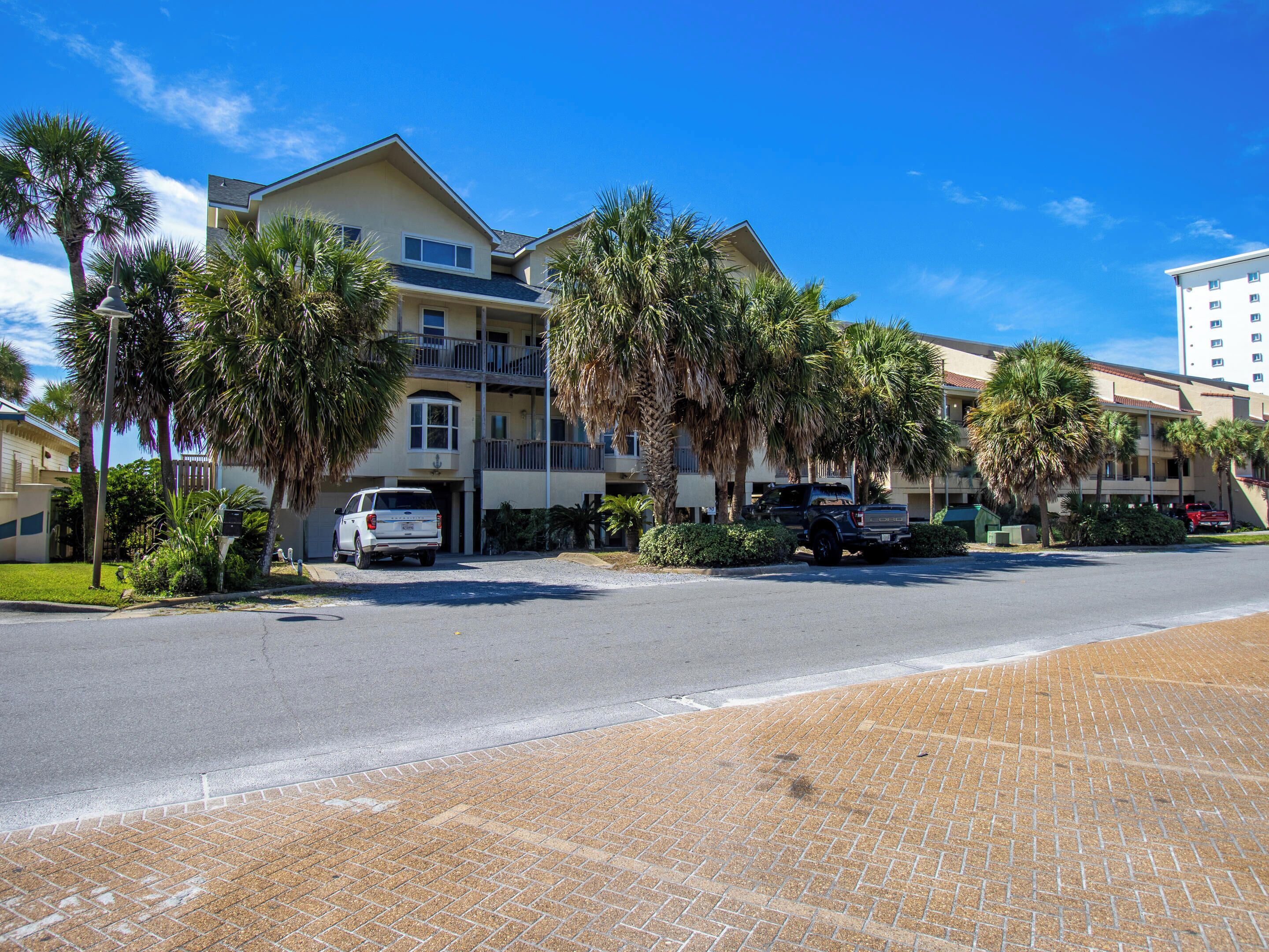 A multi-story hotel with a balcony on the second floor, a parking area with several cars, and palm trees in the foreground.