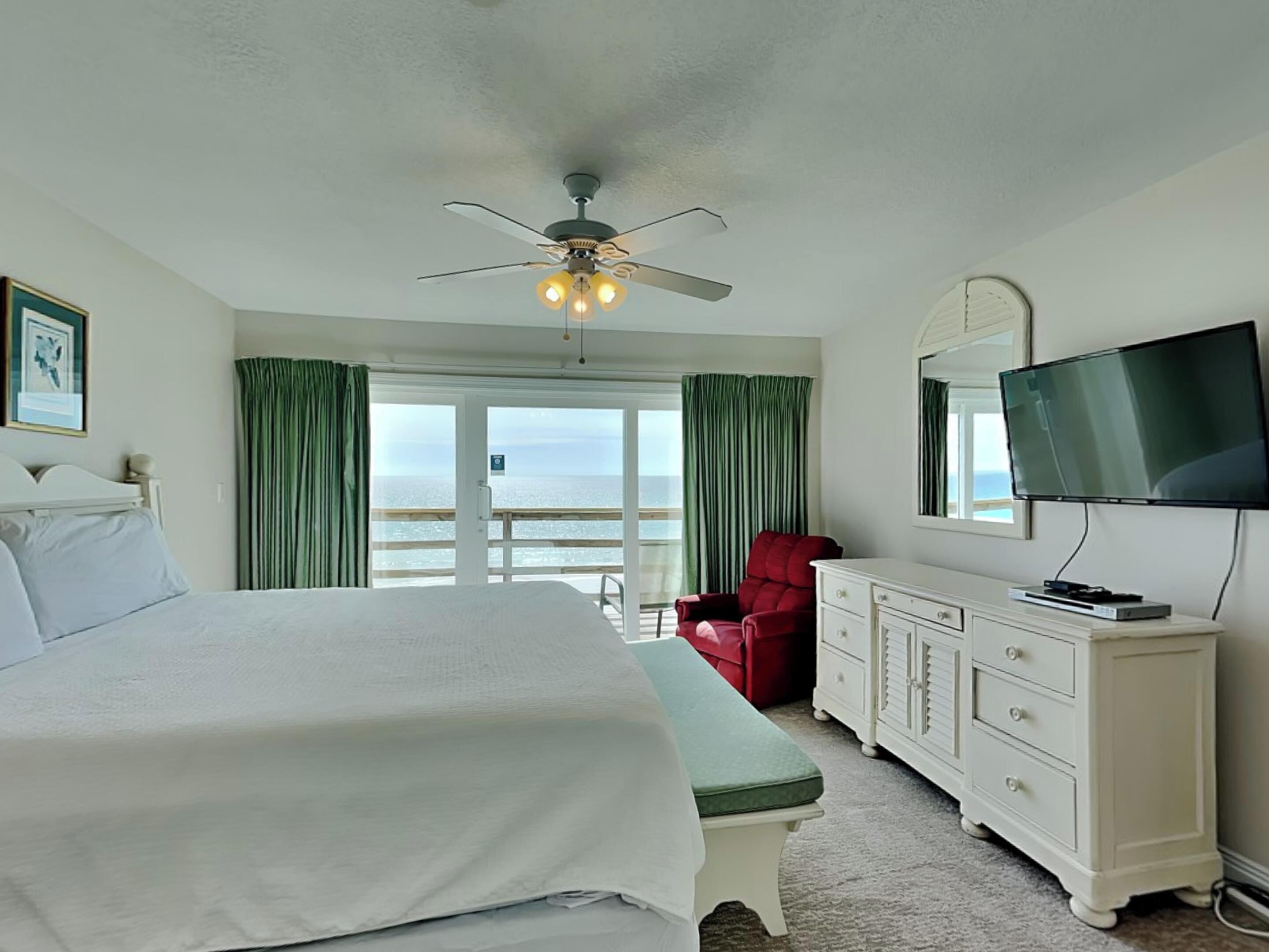 A hotel room with a bed, a red chair, a television, and a balcony with a view of the sea.