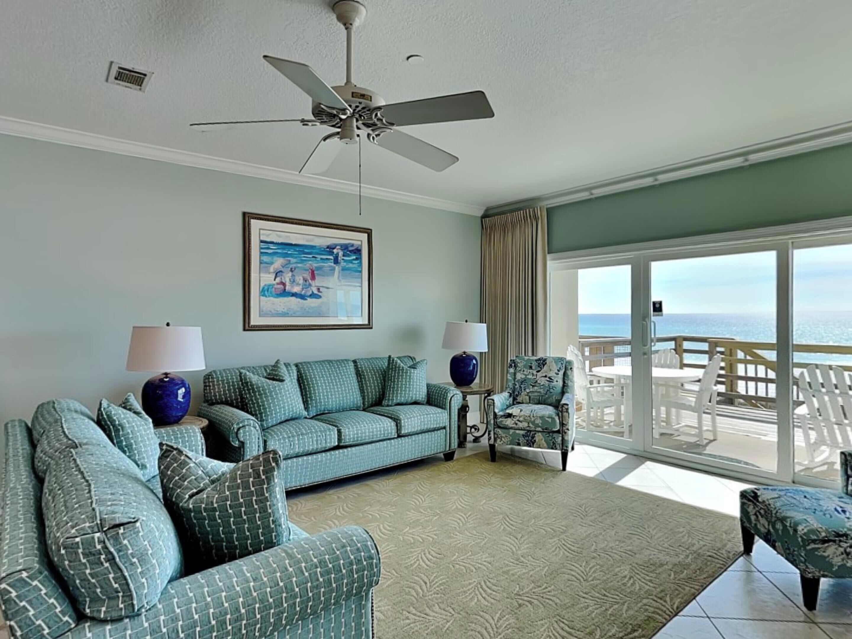A living room with a sofa, chairs, a ceiling fan, a painting, and a view of the ocean.