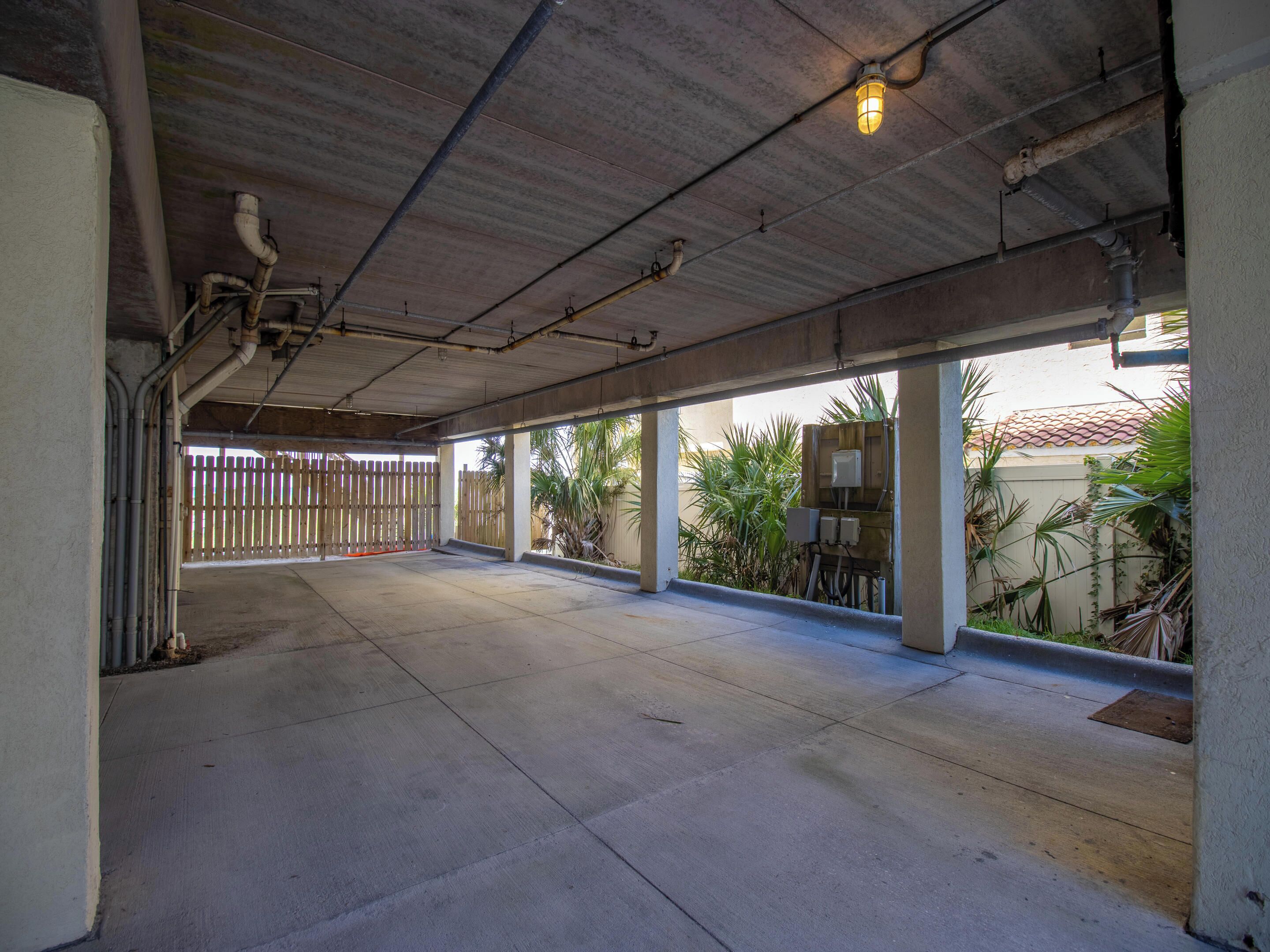 An empty covered parking area with concrete flooring, metal beams, and a wooden fence in the background.