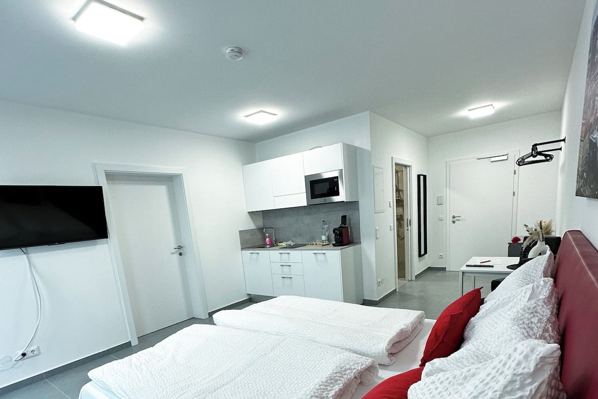 A modern hotel room with a large bed, a desk, and a kitchenette.