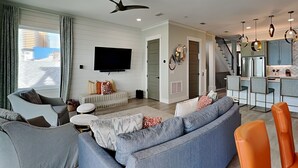 A modern living room with a grey sectional sofa, a grey armchair, and an orange chair. A flat-screen TV is mounted on the wall, and there is a staircase leading to the kitchen area. - Breezy Beach by Resort Collection (Panama City Beach)