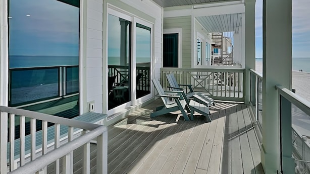 A covered deck with a view of the ocean, featuring a railing, a seating area with chairs, and a glass door leading to an outdoor space. - Breezy Beach by Resort Collection (Panama City Beach)