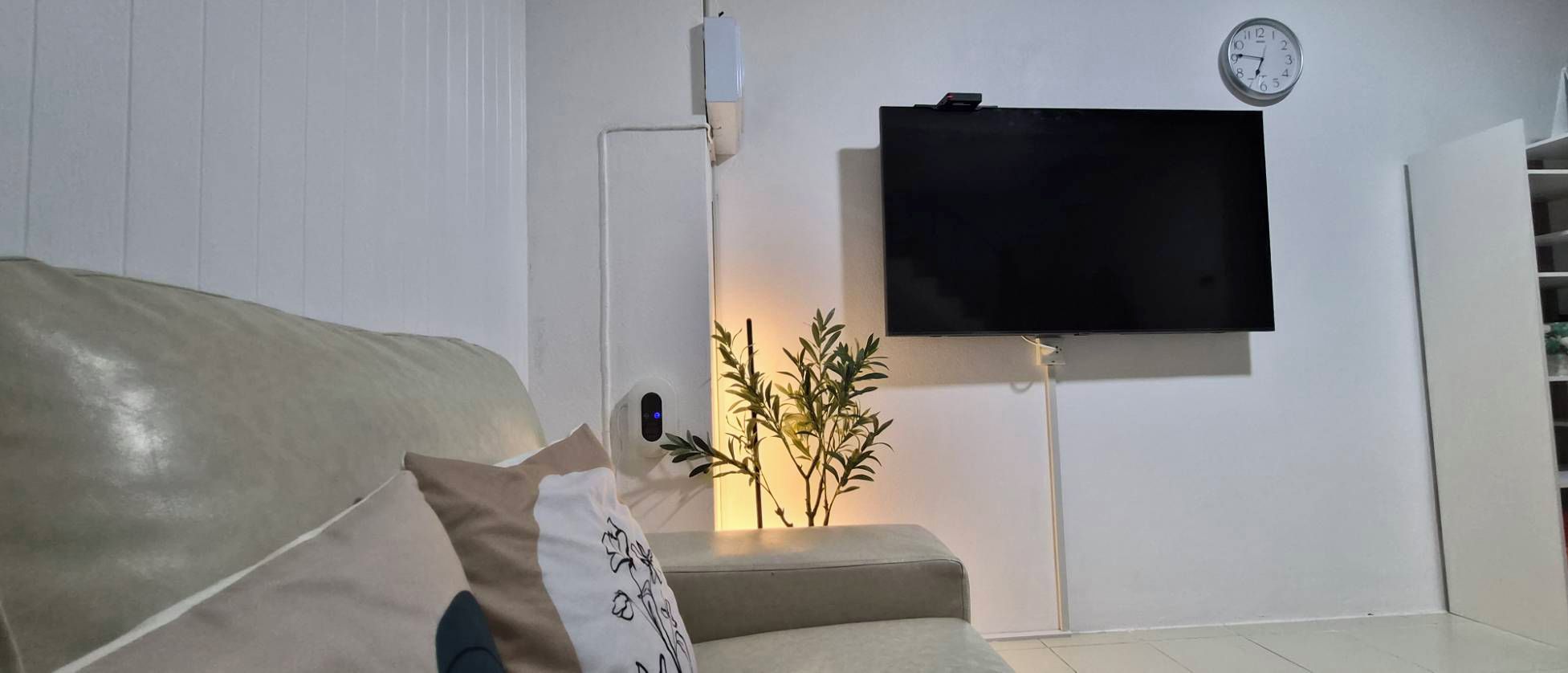 Ett vardagsrum med en soffa, en platt-TV som är monterad på väggen, en klocka och en växt.