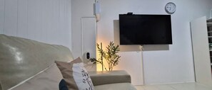 A living room with a sofa, a flat-screen TV mounted on the wall, a clock, and a plant. - Ban Sawasdee, 3 Bedrooms Townhouse, Large home, near Don Mueang Airport (Lamlukka)