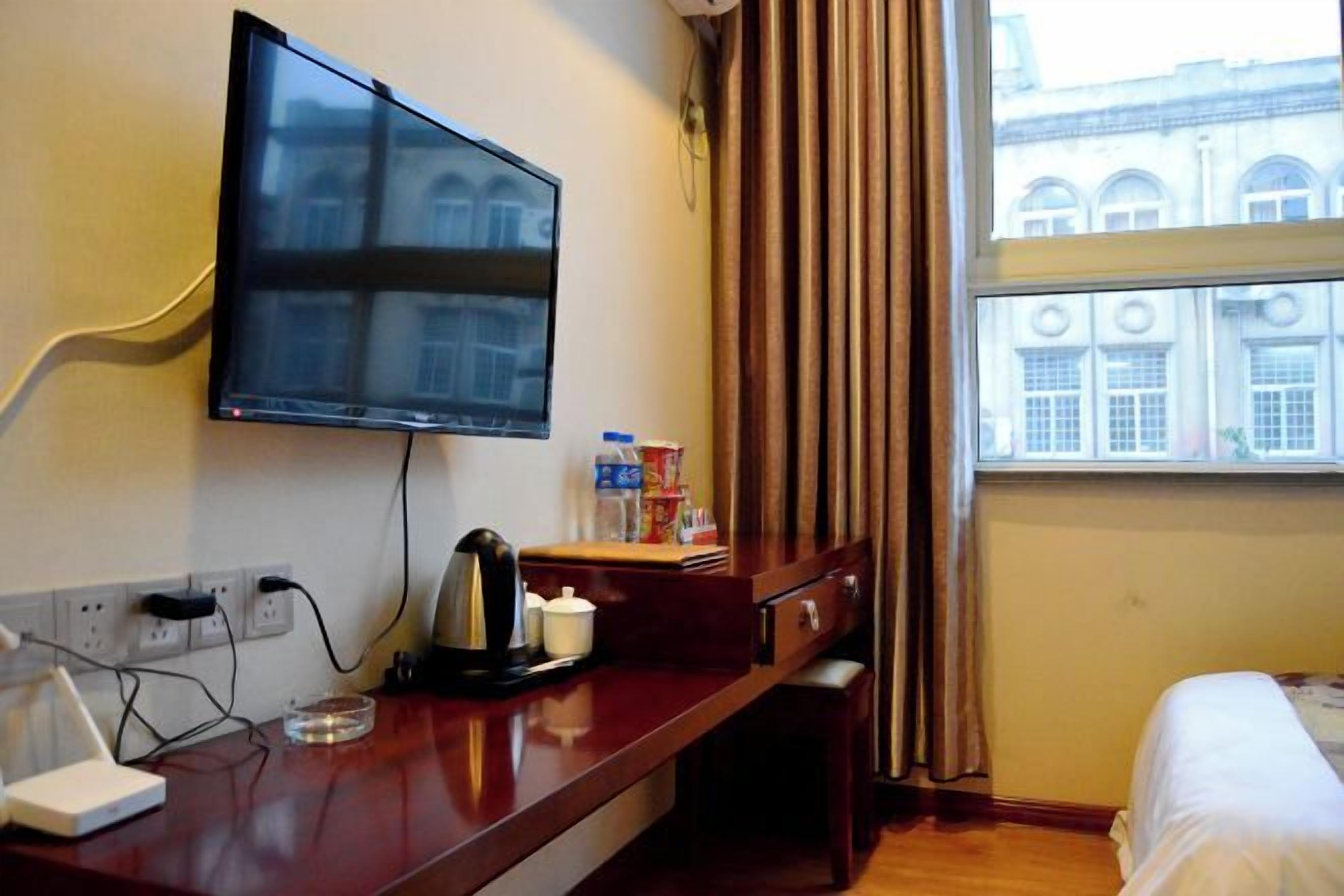 A hotel room with a flat-screen TV mounted on the wall, a wooden desk with a kettle, a cup, and a bottle of water, a bed with white bedding, and a window with curtains.