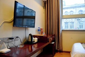Une chambre d’hôtel équipée d’un téléviseur à écran plat fixé au mur, d’un bureau en bois avec une bouilloire, une tasse et une bouteille d’eau, d’un lit avec du linge de lit blanc et d’une fenêtre avec des rideaux.