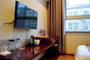 A hotel room with a flat-screen TV mounted on the wall, a wooden desk with a kettle, a cup, and a bottle of water, a bed with white bedding, and a window with curtains.