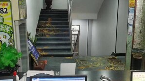 Un escalier recouvert d’une moquette à motifs menant à un étage supérieur, un bureau avec un ordinateur, et divers panneaux et affiches accrochés aux murs.