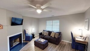 A living room with a ceiling fan, a fireplace, a television, a couch, and a desk with a lamp. - Cozy, Coastal Retreat! Minutes from Parris Island! (Port Royal)