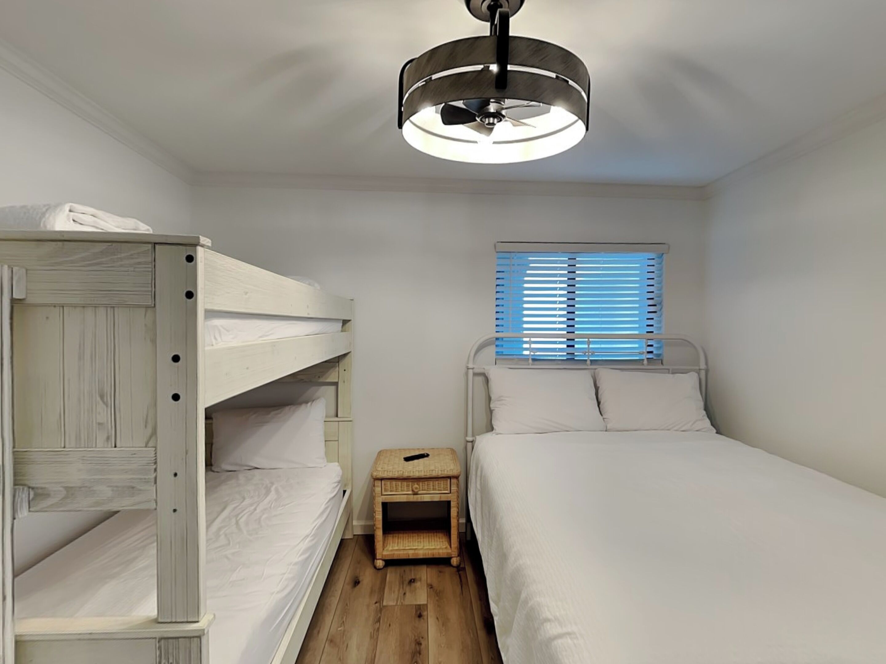 A small, clean room with bunk beds, a bedside table, and a window with blinds.