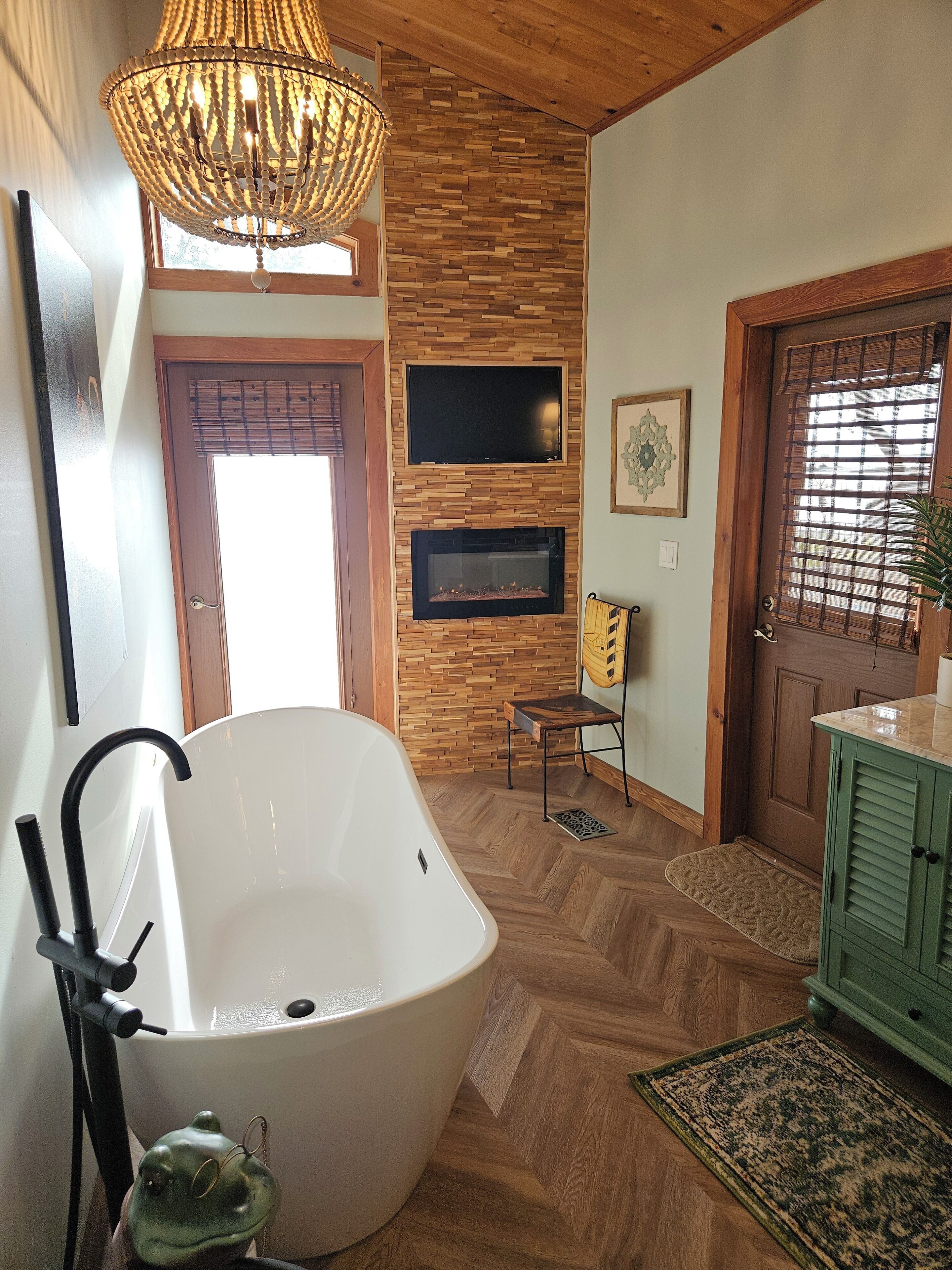 Une salle de bain avec une baignoire sur pieds, un mur en bois intégrant un foyer, un téléviseur fixé au mur et une grande fenêtre avec des stores.