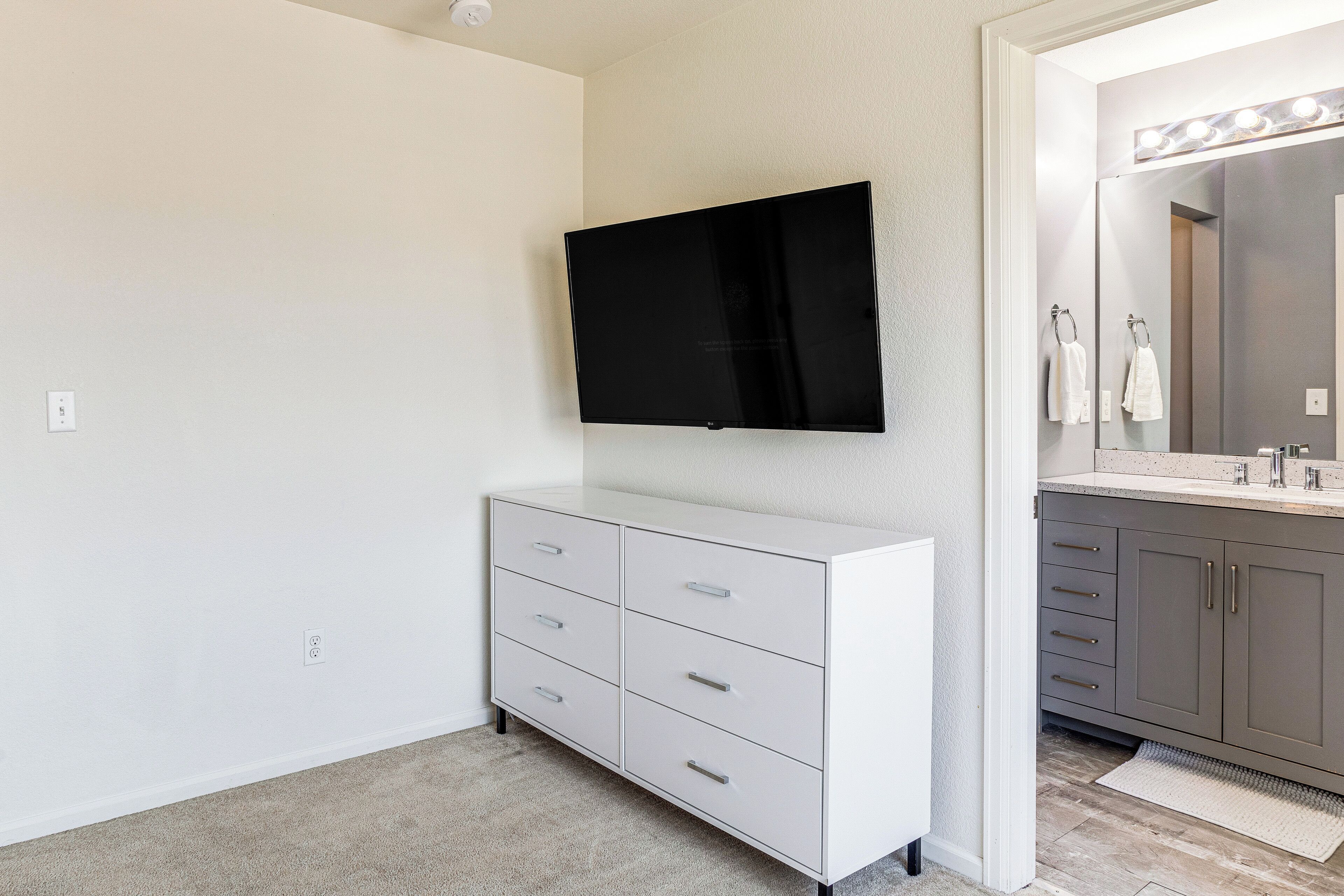 A bathroom with a white vanity, a mirror with lights, and a flat-screen TV mounted above the vanity.