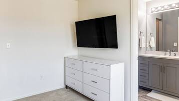 A bathroom with a white vanity, a mirror with lights, and a flat-screen TV mounted above the vanity.
