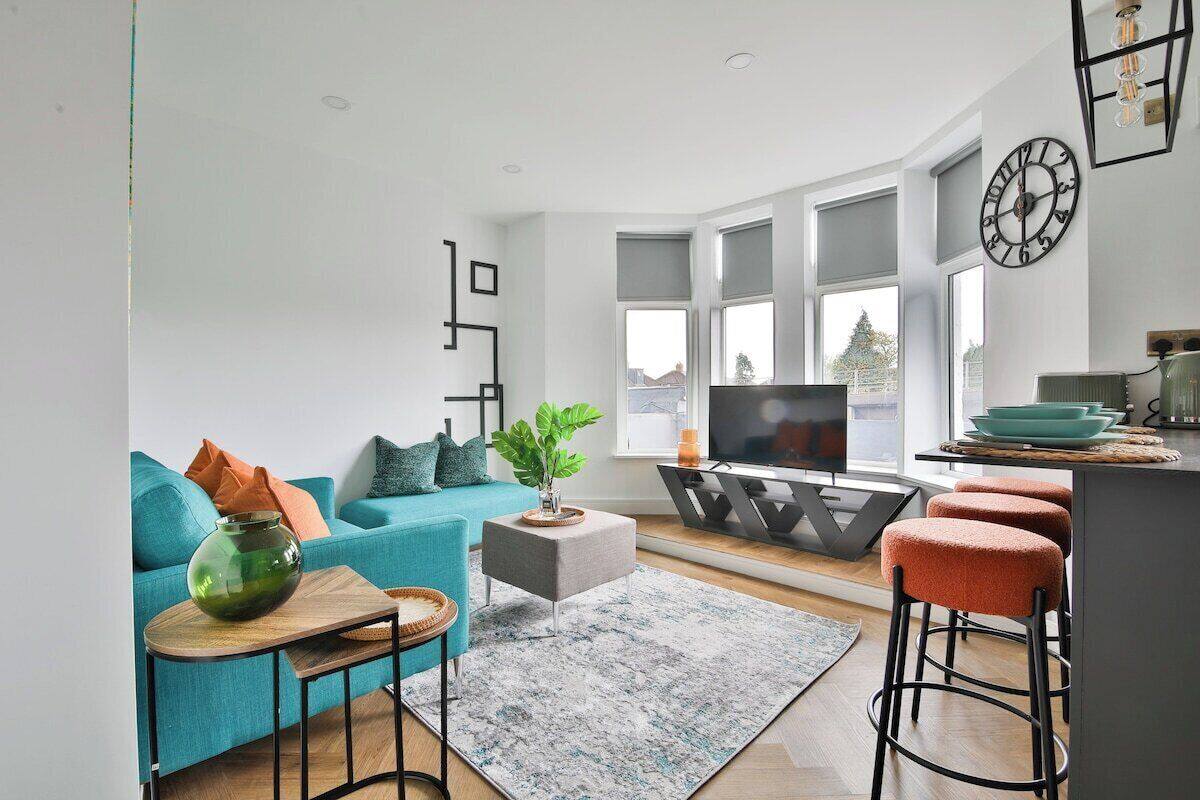 A modern living room with a teal sofa, a coffee table, a TV, and a kitchen area with stools.