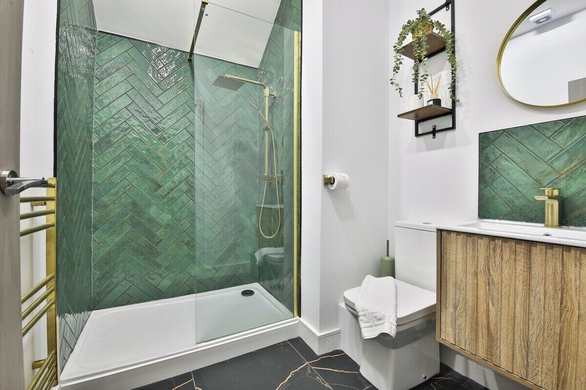 A modern bathroom with a glass shower enclosure, a wooden vanity, and a green herringbone tile backsplash.