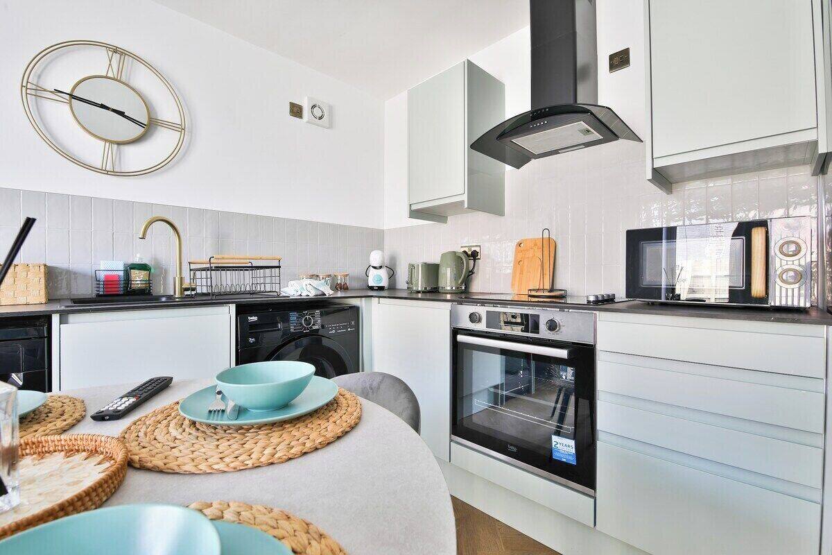 A modern kitchen with white cabinets, a built-in oven, and a microwave. There is a clock on the wall and a sink with a faucet.