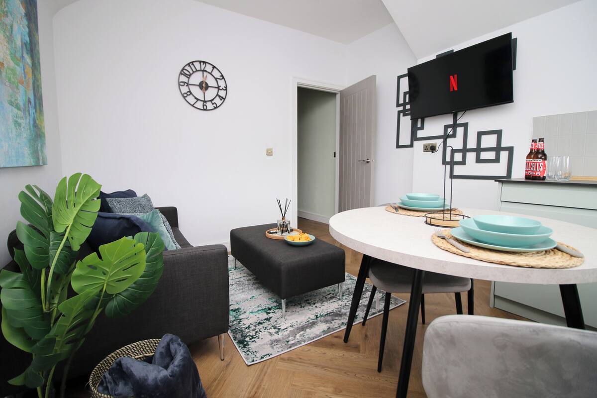 A modern living room with a sofa, ottoman, and a dining table with blue plates. A flat-screen TV is mounted on the wall, and a clock is visible.