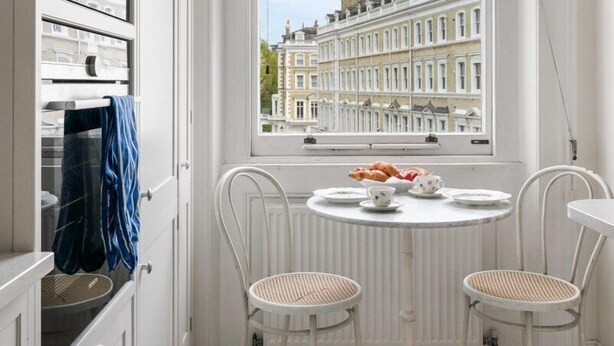 A kitchen with white cabinetry, a window with a view of terraced houses, and a small round table with two chairs. - Exceptional 2 bed, 2 bath, South Ken Apt (London)