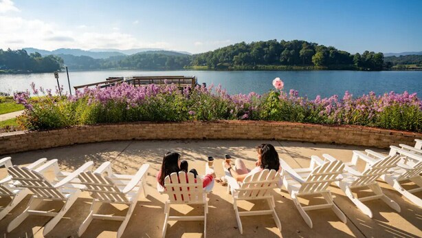 A lakeside seating area with white chairs and a wooden deck overlooking a lake, surrounded by purple flowers and a brick wall. - Biltmore discount tickets|close to Cataloochee ski (Waynesville)